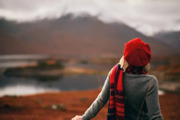 A woman wearing a red beret and scarf gazes at a serene autumn landscape by a lake, with distant mountains in the background. What Makes a Scottish Scarf the Perfect Accessory for Any Season?